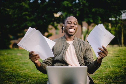 A masculine-presenting person of color showing a big smile while sitting on a green lawn holding papers in both hands and a laptop on their lap.The person’s expression of glee is what I hope students in my courses feel about learning!